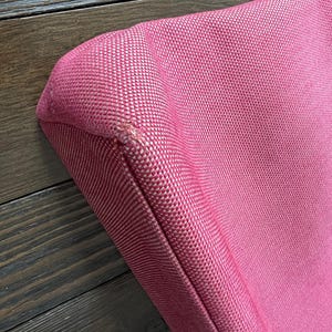 May include: A close-up of a pink tote bag with a textured fabric. The bag has black leather handles and a small rectangular metal emblem. The bag's corner shows a slight imperfection in the stitching.