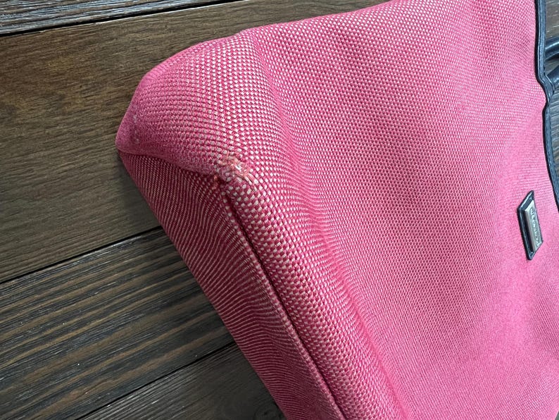 May include: A close-up of a pink tote bag with a textured fabric. The bag has black leather handles and a small rectangular metal emblem. The bag's corner shows a slight imperfection in the stitching.