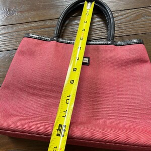 May include: A red tote bag with black handles and trim. The bag is approximately 12 inches long. The bag has a rectangular shape and a small label on the front. The bag is made of a textured material.