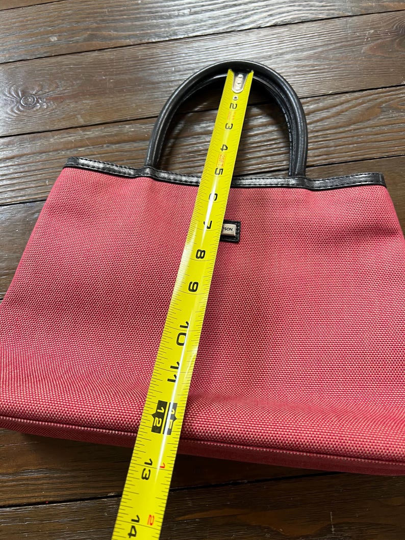 May include: A red tote bag with black handles and trim. The bag is approximately 12 inches long. The bag has a rectangular shape and a small label on the front. The bag is made of a textured material.