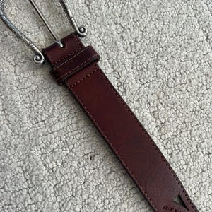 Peut inclure: Une ceinture en cuir marron avec une boucle argent&eacute;e. La boucle pr&eacute;sente un motif orn&eacute;. La ceinture comporte des &eacute;l&eacute;ments d&eacute;coratifs d&eacute;coup&eacute;s pr&egrave;s de l'extr&eacute;mit&eacute;. La ceinture mesure environ 3,8 cm de large.