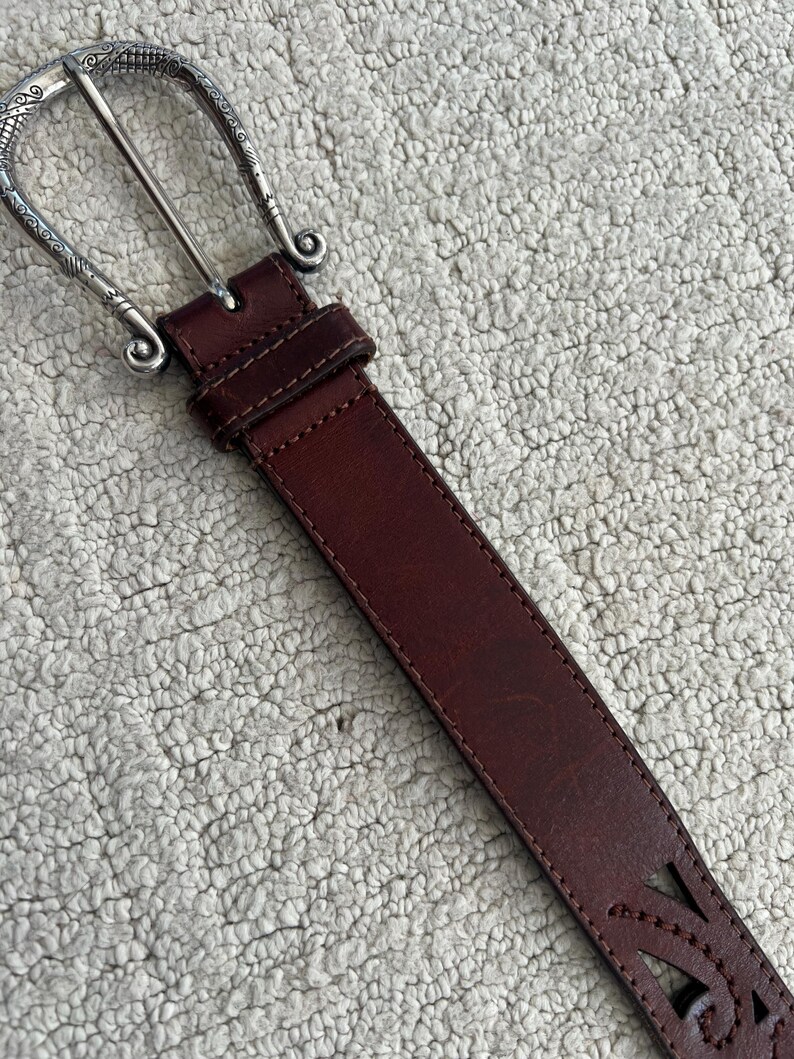 Peut inclure: Une ceinture en cuir marron avec une boucle argent&eacute;e. La boucle pr&eacute;sente un motif orn&eacute;. La ceinture comporte des &eacute;l&eacute;ments d&eacute;coratifs d&eacute;coup&eacute;s pr&egrave;s de l'extr&eacute;mit&eacute;. La ceinture mesure environ 3,8 cm de large.