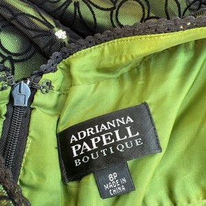 May include: Close-up of a green dress with black floral lace overlay. The dress features a zipper and a tag that reads "Adrianna Papell Boutique" and "8P Made in China". The dress has a black lace trim.