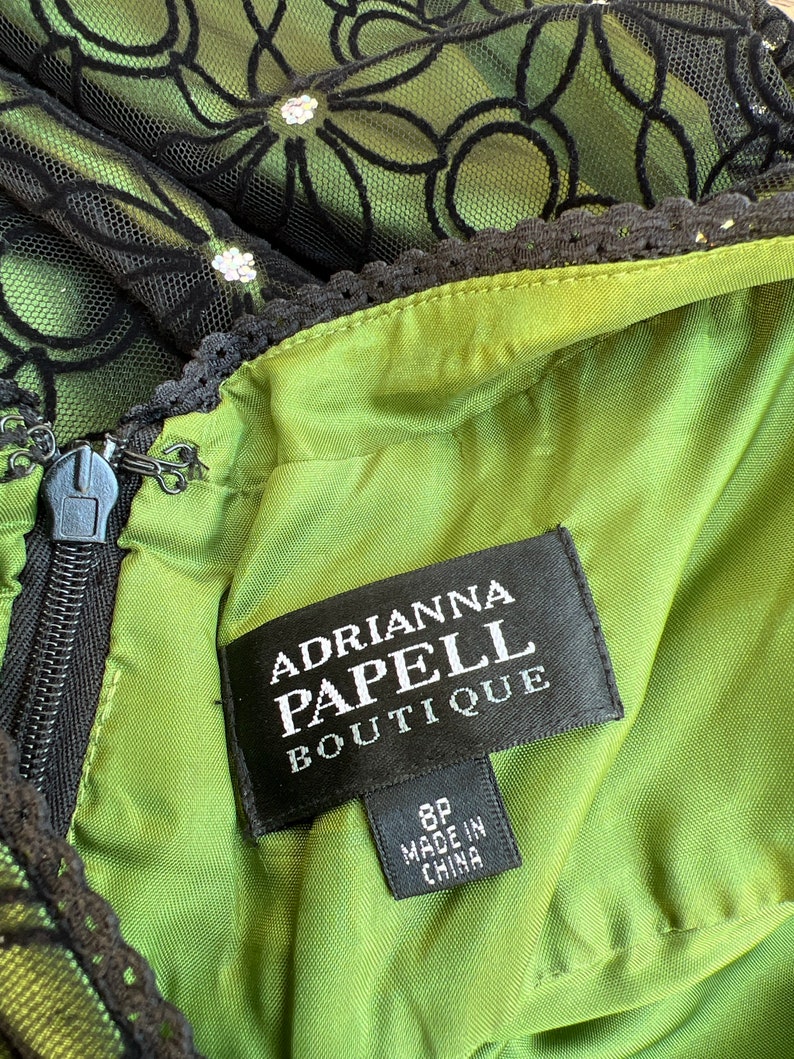 May include: Close-up of a green dress with black floral lace overlay. The dress features a zipper and a tag that reads "Adrianna Papell Boutique" and "8P Made in China". The dress has a black lace trim.