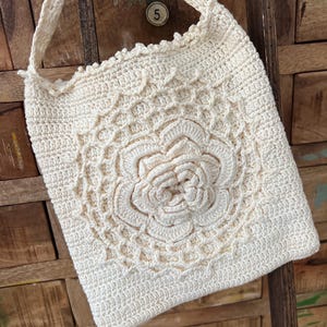 May include: Cream-colored crochet tote bag with a floral design on the front. The bag features a long strap and is made of a textured yarn. The floral design is raised, adding dimension to the bag.