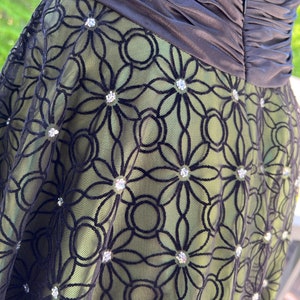 May include: A formal dress featuring a black bodice and a green skirt overlaid with black floral lace. The lace is embellished with sparkling rhinestones. The dress has a ruched bodice and a zipper closure.