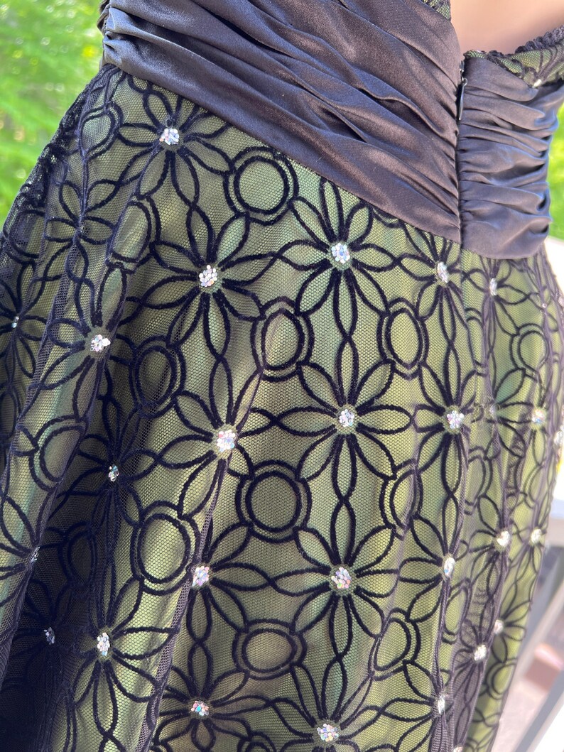 May include: A formal dress featuring a black bodice and a green skirt overlaid with black floral lace. The lace is embellished with sparkling rhinestones. The dress has a ruched bodice and a zipper closure.
