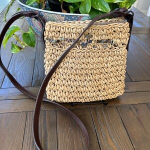 May include: A woven straw crossbody bag with a brown leather strap. The bag has a rectangular shape with a zipper closure and a decorative beaded detail. The word "SCALA" is visible on the bag. The bag is sitting on a wooden surface.