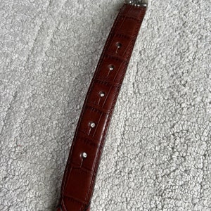 May include: A brown leather belt with a braided design. The belt has a textured, crocodile-like pattern on the main strap and a decorative silver buckle. The belt has several holes for adjustment.