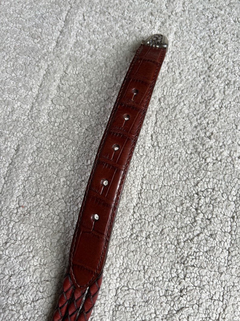 May include: A brown leather belt with a braided design. The belt has a textured, crocodile-like pattern on the main strap and a decorative silver buckle. The belt has several holes for adjustment.
