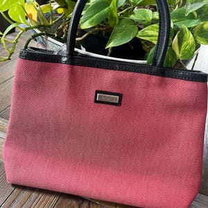 May include: A small, rectangular, red handbag with black leather handles and trim. The bag has a silver rectangular nameplate that reads "JIM THOMPSON". The bag is sitting on a wooden surface.