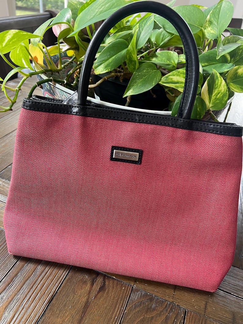 May include: A small, rectangular, red handbag with black leather handles and trim. The bag has a silver rectangular nameplate that reads "JIM THOMPSON". The bag is sitting on a wooden surface.