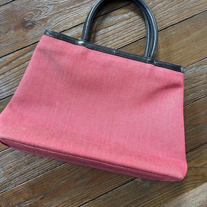 May include: A pink tote bag with black leather handles and trim. The bag is made of a textured fabric and has a rectangular shape. The handles are curved and attached to the top of the bag.