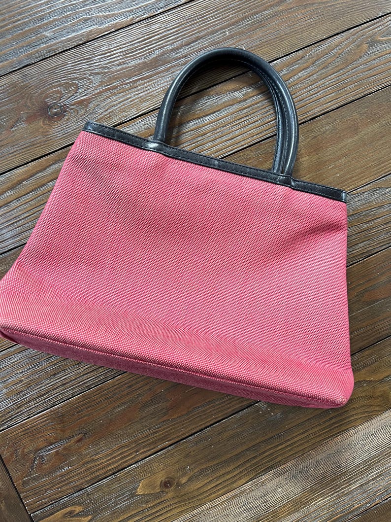 May include: A pink tote bag with black leather handles and trim. The bag is made of a textured fabric and has a rectangular shape. The handles are curved and attached to the top of the bag.