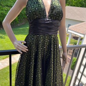 May include: A green halter dress with a black floral lace overlay. The dress features a black satin waistband and is adorned with sparkling embellishments. The neckline is trimmed with black beading. The dress is displayed on a mannequin.