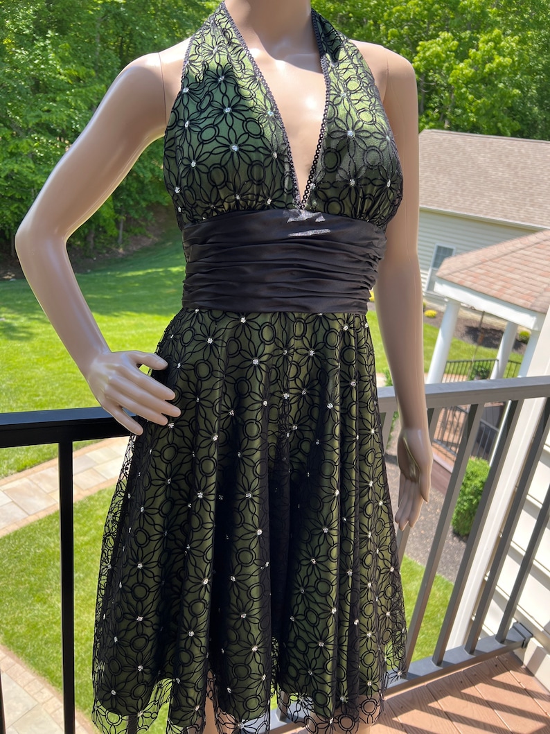 May include: A green halter dress with a black floral lace overlay. The dress features a black satin waistband and is adorned with sparkling embellishments. The neckline is trimmed with black beading. The dress is displayed on a mannequin.