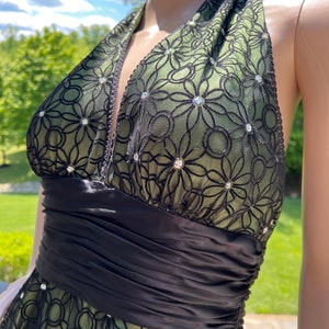 May include: A halter-style dress featuring a green underlay with a black floral lace overlay. The dress has a black satin waistband and is embellished with sparkling rhinestones. The dress is designed for formal occasions.