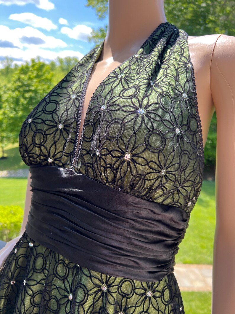 May include: A halter-style dress featuring a green underlay with a black floral lace overlay. The dress has a black satin waistband and is embellished with sparkling rhinestones. The dress is designed for formal occasions.