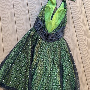 May include: A green halter dress with a black floral overlay and a black satin waist. The dress features a low back and a bow at the neck. The overlay is embellished with small, white, round accents.