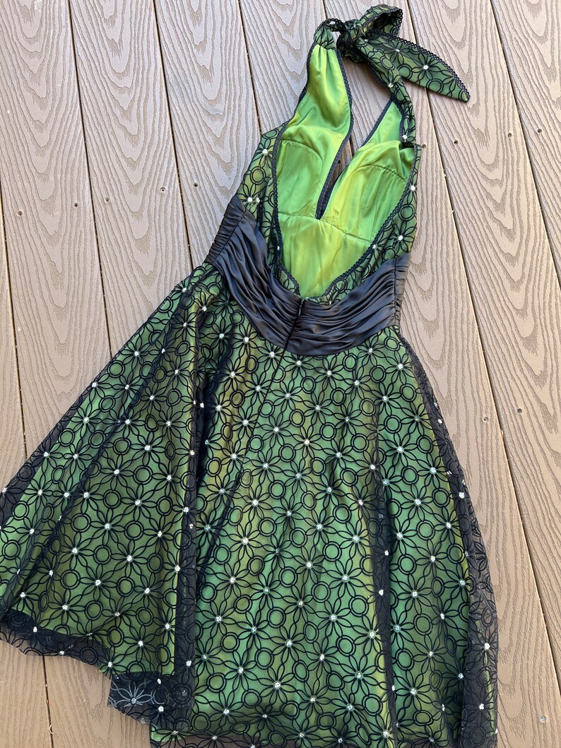 May include: A green halter dress with a black floral overlay and a black satin waist. The dress features a low back and a bow at the neck. The overlay is embellished with small, white, round accents.