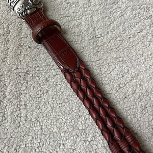 May include: A brown leather belt with a silver buckle. The belt features a braided design and a textured, crocodile-like pattern. The silver buckle has an ornate, detailed design.