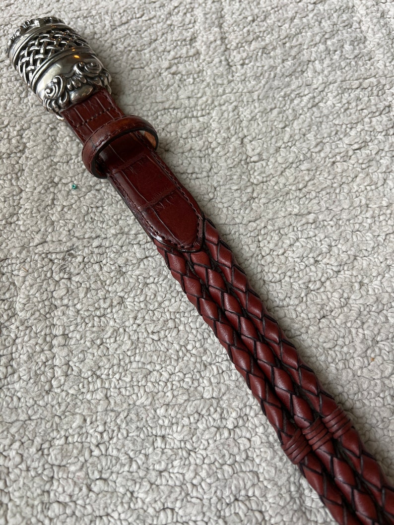 May include: A brown leather belt with a silver buckle. The belt features a braided design and a textured, crocodile-like pattern. The silver buckle has an ornate, detailed design.