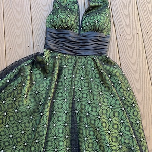 May include: A green halter dress with a black floral overlay. The dress features a black satin waistband and a matching halter tie. The skirt has a flared silhouette and is adorned with small, sparkling accents.