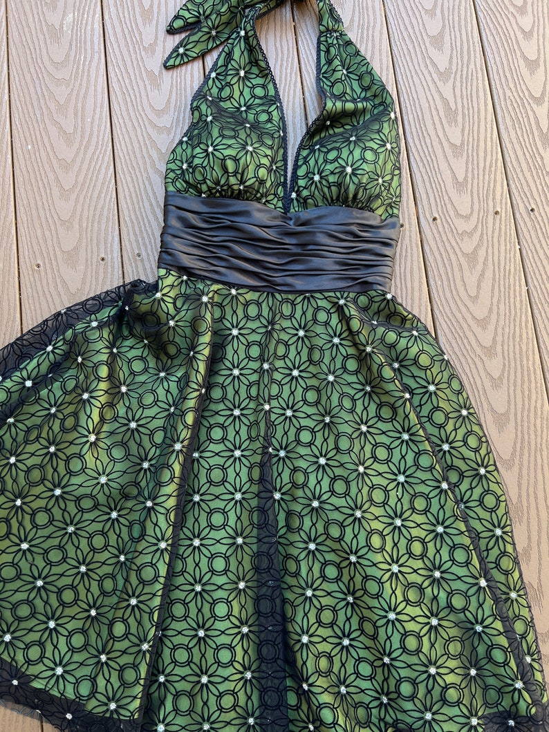 May include: A green halter dress with a black floral overlay. The dress features a black satin waistband and a matching halter tie. The skirt has a flared silhouette and is adorned with small, sparkling accents.
