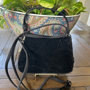 May include: Black velvet handbag with a decorative swirl pattern. The bag has a gold-colored clasp and a long, adjustable black strap. The bag also has a short handle. The bag is sitting on a wooden surface.
