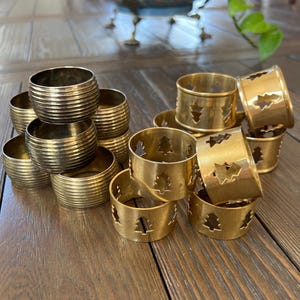 Puede incluir: Un conjunto de servilleteros de latón. Algunos anillos tienen un diseño acanalado, mientras que otros presentan un diseño de árbol de Navidad recortado. Los anillos están dispuestos sobre una superficie de madera.