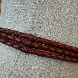 May include: Close-up of a brown leather belt with a braided design. The belt has three braided strands and is secured with a matching leather wrap. The belt is displayed against a textured, silver background.