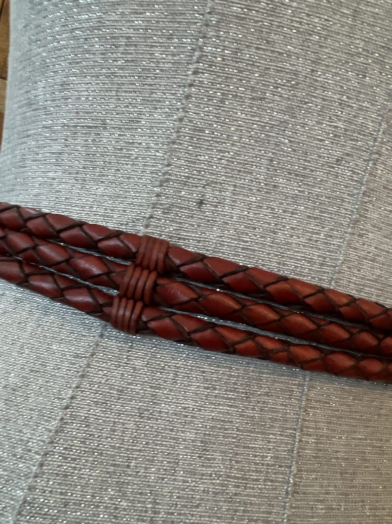 May include: Close-up of a brown leather belt with a braided design. The belt has three braided strands and is secured with a matching leather wrap. The belt is displayed against a textured, silver background.