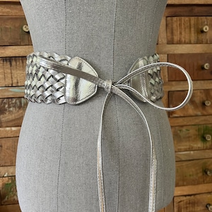 May include: A silver-coloured, wide belt with a woven design and a bow closure. The belt is made of a shiny, metallic material and is displayed on a mannequin. The belt's width is approximately 7.6 cm.