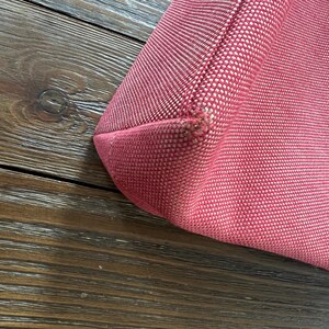 May include: Close-up of a red tote bag corner. The bag is made of a textured fabric with a woven pattern. The corner shows a seam and slight wear. The background is a dark wood surface.