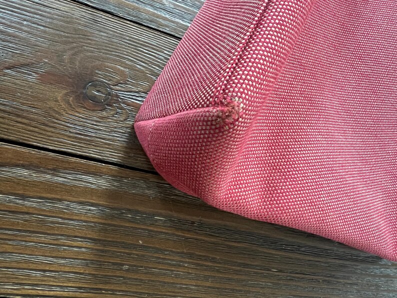 May include: Close-up of a red tote bag corner. The bag is made of a textured fabric with a woven pattern. The corner shows a seam and slight wear. The background is a dark wood surface.