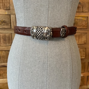 May include: A brown leather belt with a silver-coloured buckle. The buckle has a woven centre design and ornate detailing. The belt is displayed on a mannequin. The belt is approximately 3.2 cm wide.