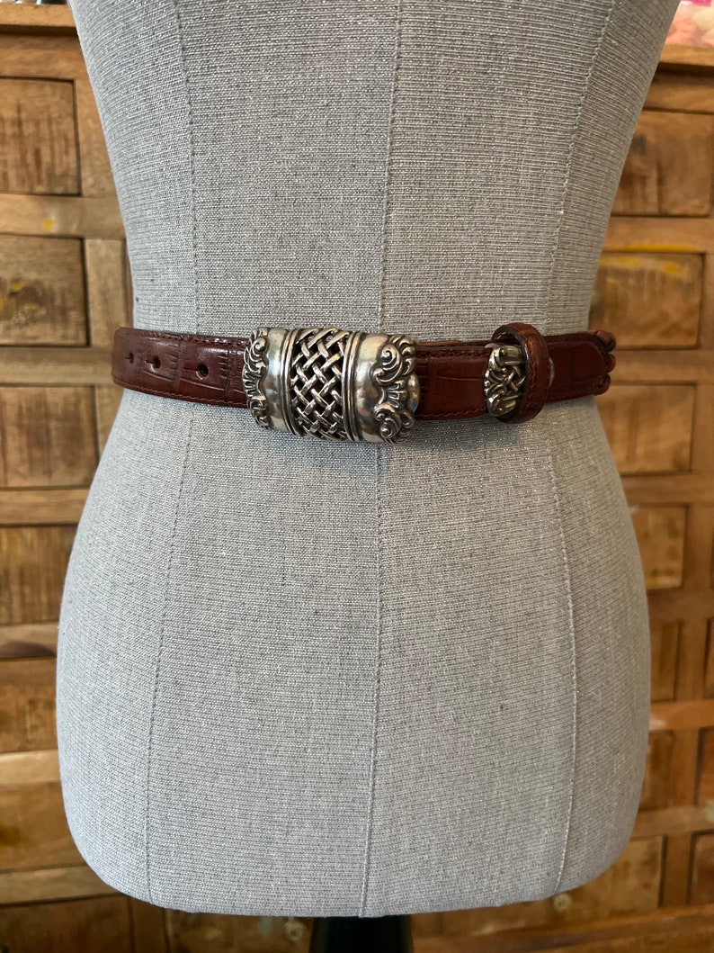 May include: A brown leather belt with a silver-coloured buckle. The buckle has a woven centre design and ornate detailing. The belt is displayed on a mannequin. The belt is approximately 3.2 cm wide.