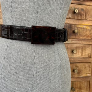 May include: A dark brown belt with a rectangular buckle. The belt has a textured, crocodile-like pattern. The buckle is a tortoiseshell color. The belt is displayed on a mannequin.
