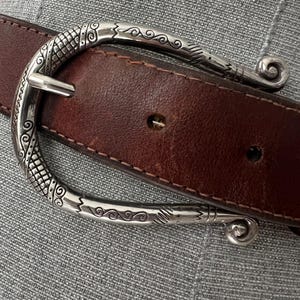 Peut inclure: Une ceinture en cuir marron avec une boucle d&eacute;corative argent&eacute;e. La boucle pr&eacute;sente un motif orn&eacute; avec des motifs tourbillonnants et une fermeture &agrave; ardillon classique. La ceinture est cousue avec un fil contrastant.