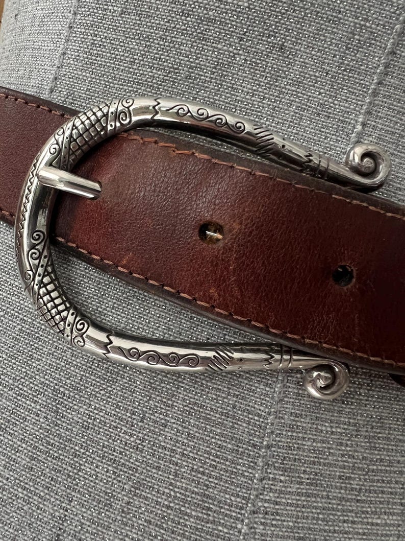 Peut inclure: Une ceinture en cuir marron avec une boucle d&eacute;corative argent&eacute;e. La boucle pr&eacute;sente un motif orn&eacute; avec des motifs tourbillonnants et une fermeture &agrave; ardillon classique. La ceinture est cousue avec un fil contrastant.