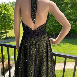May include: A halter-neck dress with a black and olive green color scheme. The dress features a black bodice and a skirt with a floral lace overlay. The halter strap and the lace are embellished with small, white accents.