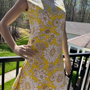 May include: A sleeveless yellow dress with a white floral pattern and white lace trim. The dress has a round neckline. The background shows a green lawn and trees. The dress is displayed on a mannequin.