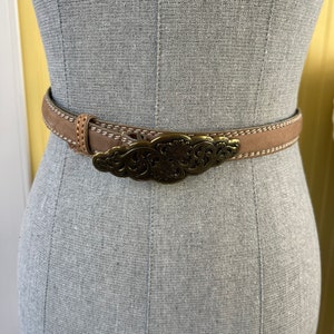 Puede incluir: Un cinturón color canela con una hebilla de latón antiguo. El cinturón marrón claro tiene costuras blancas a lo largo de los bordes. La hebilla presenta un diseño floral ornamentado. El cinturón se muestra en un maniquí.