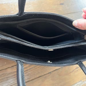 May include: Black leather tote bag with two handles. The bag has a zippered top and multiple interior compartments. The interior is black. The bag is open, revealing the inside.