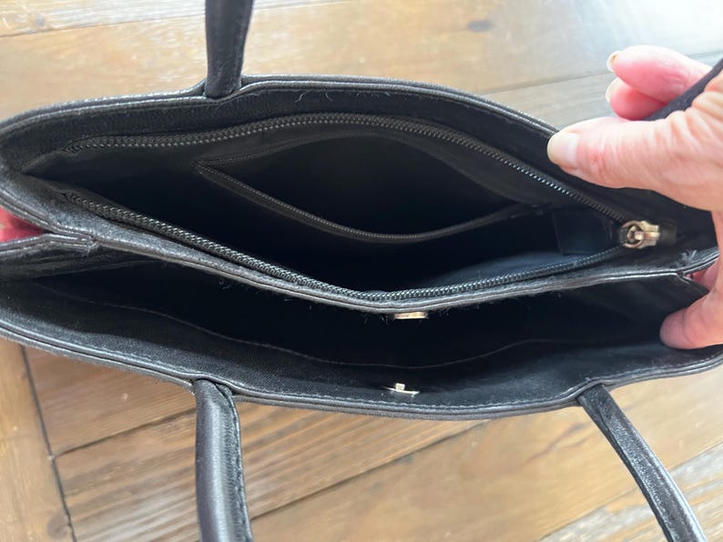 May include: Black leather tote bag with two handles. The bag has a zippered top and multiple interior compartments. The interior is black. The bag is open, revealing the inside.
