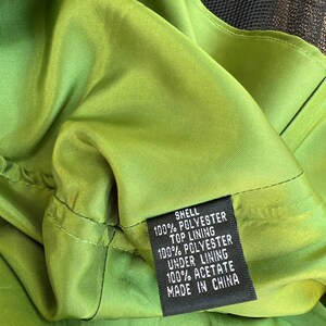 May include: Close-up of a green garment's interior, showcasing the fabric's texture and stitching. A black tag displays the composition: shell 100% polyester, top lining 100% polyester, under lining 100% acetate. The garment is made in China.