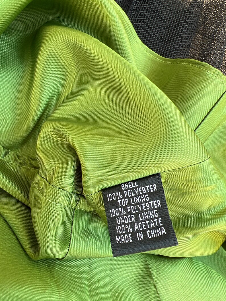 May include: Close-up of a green garment's interior, showcasing the fabric's texture and stitching. A black tag displays the composition: shell 100% polyester, top lining 100% polyester, under lining 100% acetate. The garment is made in China.
