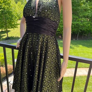 May include: A halter-neck dress with a black floral lace overlay over a green lining. The dress features a black satin waistband and is adorned with small, sparkling embellishments. The dress is displayed on a mannequin.