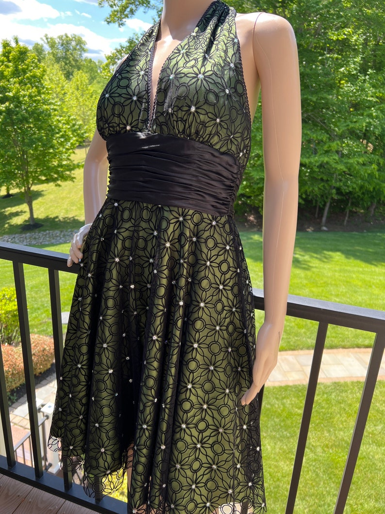 May include: A halter-neck dress with a black floral lace overlay over a green lining. The dress features a black satin waistband and is adorned with small, sparkling embellishments. The dress is displayed on a mannequin.