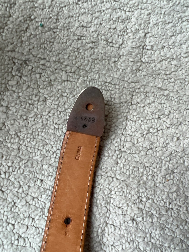 May include: Close-up of a brown leather belt with white stitching. The belt has a metal end piece with the number "1996" stamped on it. The word "CHINA" is also visible on the belt.
